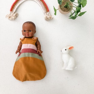 May include: A doll wearing a mustard-colored dress with pink and green stripes. A white ceramic bunny with pink ears sits nearby. A rainbow-shaped wall hanging and a small potted plant complete the scene, all against a white background.