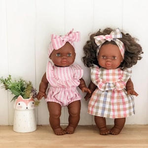 Peut inclure: Deux poupées à la peau brune et aux cheveux noirs portant des robes à rayures roses et blanches et à carreaux avec des cols à volants. Les poupées portent des bandeaux assortis à rayures roses et blanches avec des nœuds.