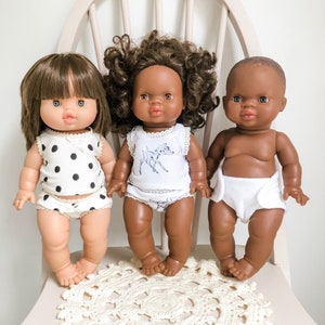 Puede incluir: Tres muñecas sentadas en una silla blanca con un mantel de encaje blanco. Las muñecas llevan diferentes atuendos. La muñeca de la izquierda lleva un top de tirantes de lunares blancos y negros y ropa interior blanca. La muñeca del medio lleva un top de tirantes blanco con estampado de ciervo y ropa interior blanca. La muñeca de la derecha lleva ropa interior blanca.