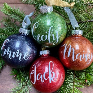 May include: Four round, glittery Christmas ornaments with names written on them in white. The ornaments are green, blue, red, and orange.