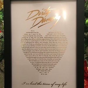 May include: A framed print of the lyrics to the song "Time of My Life" from the movie "Dirty Dancing". The lyrics are printed in gold lettering on a white background. The frame is black.