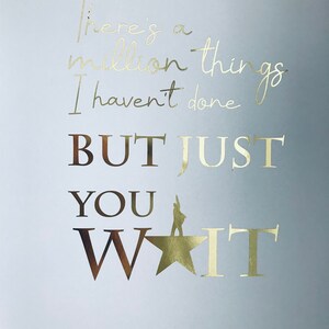 Op de afbeelding: Een lichtblauwe achtergrond met gouden folielettering die "There's a million things I haven't done BUT JUST YOU WAIT" luidt, met een gouden ster en een silhouet van een persoon die op de ster staat. De tekst "HAMILTON" staat onder de ster in gouden folielettering.