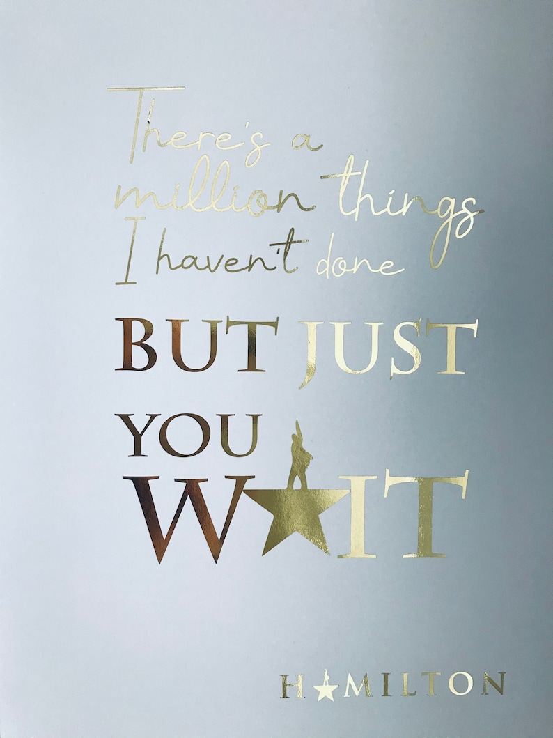Op de afbeelding: Een lichtblauwe achtergrond met gouden folielettering die "There's a million things I haven't done BUT JUST YOU WAIT" luidt, met een gouden ster en een silhouet van een persoon die op de ster staat. De tekst "HAMILTON" staat onder de ster in gouden folielettering.