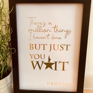 Op de afbeelding: Een ingelijst afdruk met een ontwerp in goudfolie met de tekst "There's a million things I haven't done BUT JUST YOU WAIT HAMILTON".