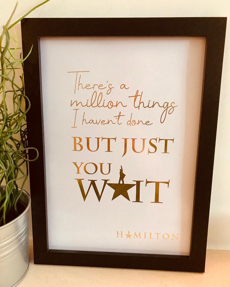 Op de afbeelding: Een ingelijst afdruk met een ontwerp in goudfolie met de tekst "There's a million things I haven't done BUT JUST YOU WAIT HAMILTON".