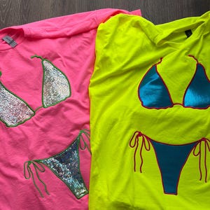 Peut inclure: Un t-shirt rose vif avec un motif de bikini holographique argenté à bordure verte et un t-shirt jaune fluo avec un motif de bikini bleu métallique à bordure rose. Les deux t-shirts ont des manches courtes et un col rond.