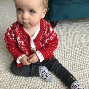 Op de afbeelding: Een baby draagt een rood-witte geruite trui met een wit onderhemd en een grijze broek. De baby draagt grijze sokken met een pingu&iuml;n-ontwerp. De baby zit op een wit, pluche tapijt.