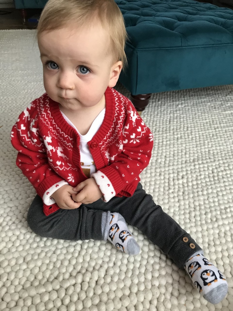 Op de afbeelding: Een baby draagt een rood-witte geruite trui met een wit onderhemd en een grijze broek. De baby draagt grijze sokken met een pingu&iuml;n-ontwerp. De baby zit op een wit, pluche tapijt.