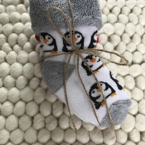 Op de afbeelding: Een paar witte babysokken met een pingu&iuml;npatroon en grijze boord. De sokken zijn met touw aan elkaar gebonden en liggen op een wit, pluche oppervlak.