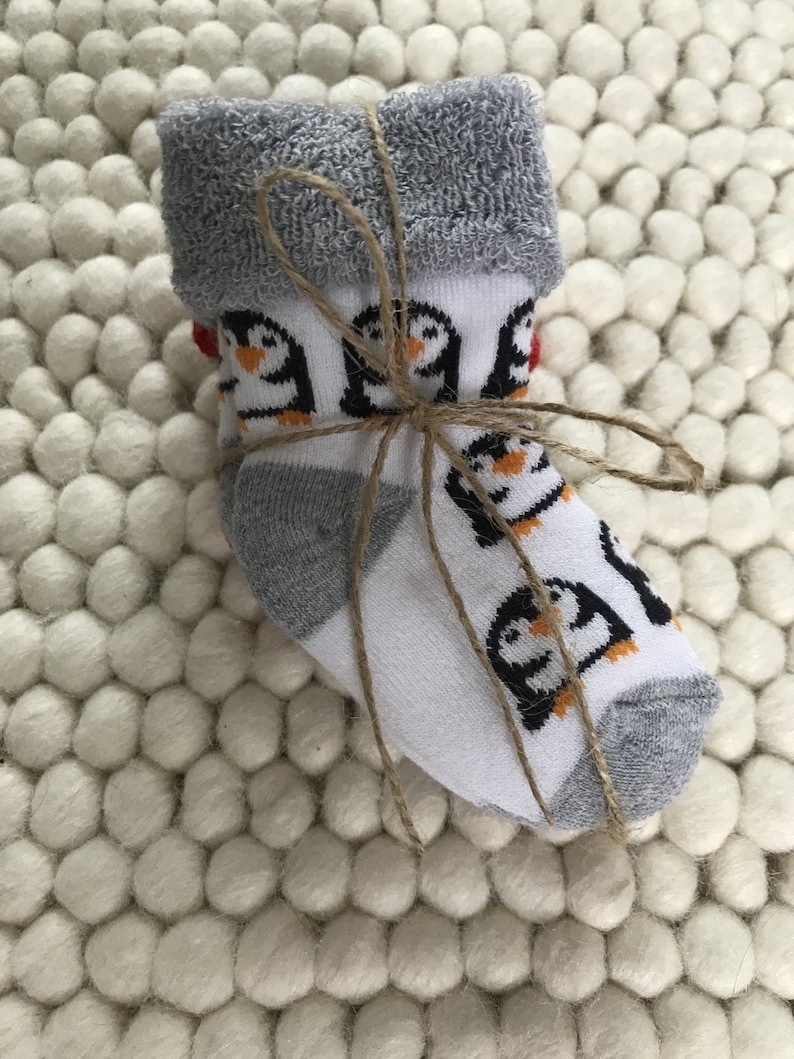 Op de afbeelding: Een paar witte babysokken met een pingu&iuml;npatroon en grijze boord. De sokken zijn met touw aan elkaar gebonden en liggen op een wit, pluche oppervlak.