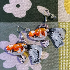 May include: A pair of gold fish earrings with silver hooks. The fish are painted with orange, white, and black details. The fish are facing opposite directions.
