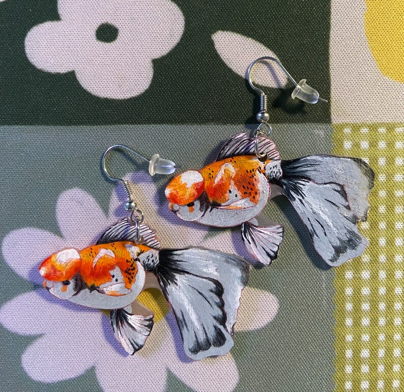 May include: A pair of gold fish earrings with silver hooks. The fish are painted with orange, white, and black details. The fish are facing opposite directions.