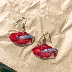 May include: A pair of silver dangle earrings with red and blue resin fish charms. The fish have a detailed painted design.