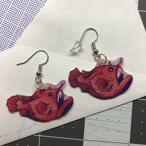 May include: A pair of dangle earrings featuring a red anglerfish with a bioluminescent lure. The earrings are made of clear acrylic with a printed image of the fish.