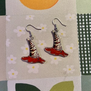 May include: A pair of earrings featuring a whimsical design of a red and white striped hat with a pointed top. The earrings are made of clear acrylic and have silver hooks.