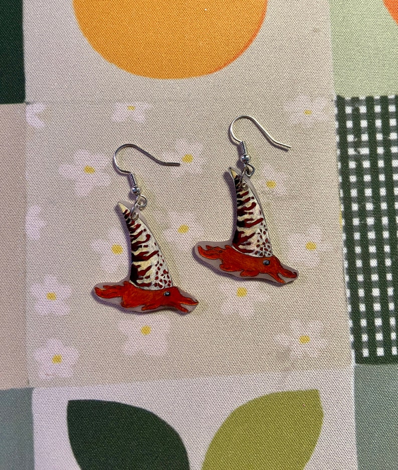 May include: A pair of earrings featuring a whimsical design of a red and white striped hat with a pointed top. The earrings are made of clear acrylic and have silver hooks.