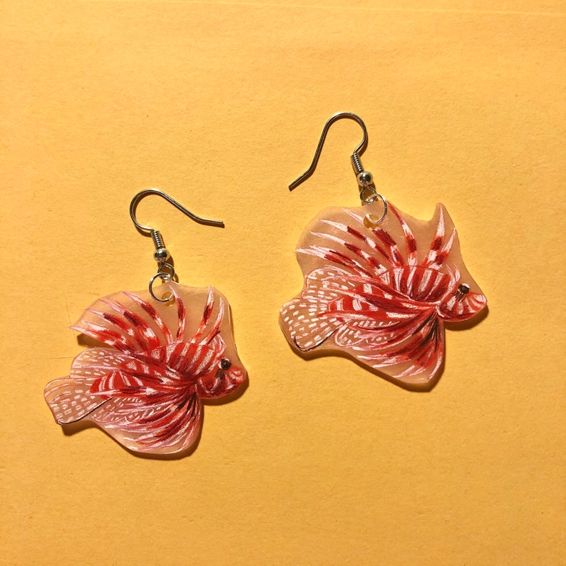 May include: A pair of earrings featuring a red and white lionfish design. The earrings are made of clear acrylic and have silver hooks.