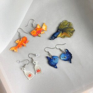 May include: A set of four pairs of dangle earrings featuring various fish designs. The earrings are made of metal and feature colorful painted fish, including a goldfish, a blue fish, and a yellow and blue fish. The earrings are perfect for adding a touch of whimsy to any outfit.