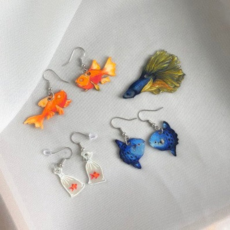May include: A set of four pairs of dangle earrings featuring various fish designs. The earrings are made of metal and feature colorful painted fish, including a goldfish, a blue fish, and a yellow and blue fish. The earrings are perfect for adding a touch of whimsy to any outfit.