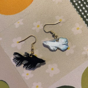 May include: A pair of dangle earrings featuring black and white fish silhouettes. The fish are painted with a watercolor effect and have gold hooks.