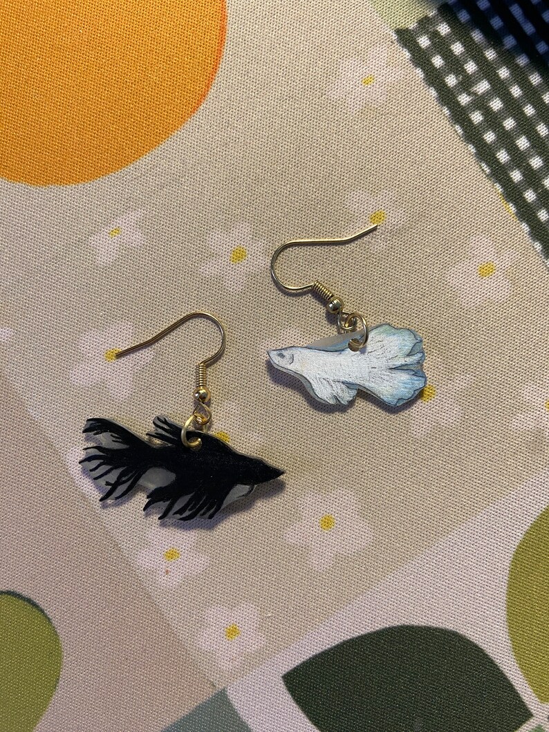 May include: A pair of dangle earrings featuring black and white fish silhouettes. The fish are painted with a watercolor effect and have gold hooks.