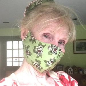 May include: A woman wearing a green floral face mask with white roses. She is wearing a red and white patterned shirt.