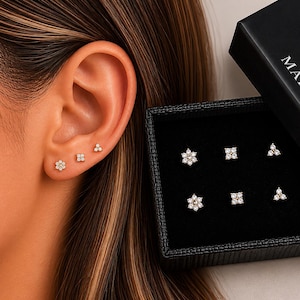 May include: A selection of six small stud earrings, presented in a black box, with three earrings worn on an ear. The earrings feature floral, square, and triangular designs, each set with multiple small, clear stones. The box is labelled "MARA".