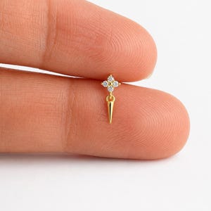 May include: A gold-tone metal earring stud featuring a four-pointed star shape adorned with four small clear round stones, with a dangling gold-tone metal spike element extending downward from the center of the star-shaped stud component.