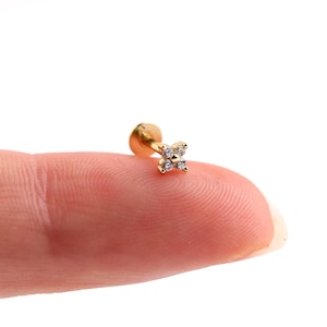 May include: A small, gold-colored earring with a four-petal flower design. The flower is adorned with small, clear gemstones. The earring is shown on a finger against a white background.