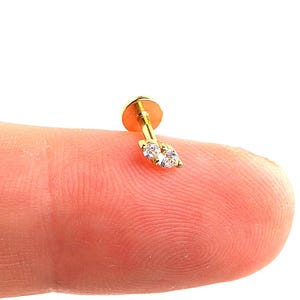 May include: A small, gold-colored earring with a flat back and a post. The earring features two clear, round gemstones set in a row. The earring is shown on a fingertip against a white background.