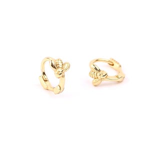 May include: Two gold-colored hoop earrings, each featuring a bee design. The earrings are small and circular, with a detailed bee charm attached to the front. The background is plain white.