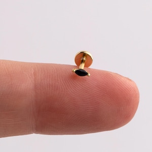Peut inclure: Une petite boucle d'oreille dorée avec une pierre noire de forme marquise. La boucle d'oreille a un dos plat et circulaire et une tige pour percer. La boucle d'oreille est présentée sur un doigt.