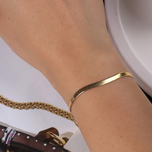 May include: A gold-toned, thin, flat, and slightly curved bracelet on a person's wrist.