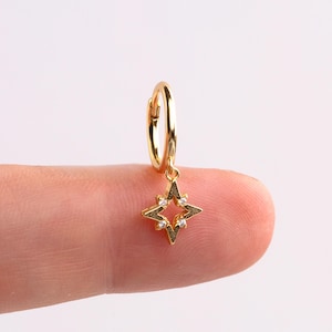 May include: A gold hoop earring with a star-shaped charm. The charm has a four-pointed design, set with small, clear gemstones. The earring is held between two fingers against a white backdrop.