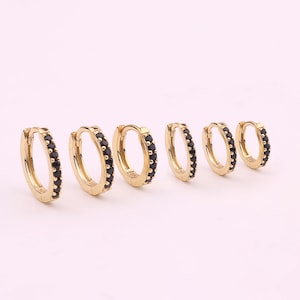 May include: Six gold-colored hoop earrings with black gemstones. The earrings are arranged in a row, showcasing varying sizes. The gemstones are set along the outer edge of each hoop, creating a sparkling effect. The earrings are displayed against a soft pink background.
