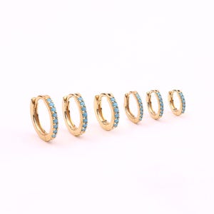 May include: A set of six gold-colored hoop earrings, each adorned with small, light blue gemstones. The earrings vary in size, with the largest on the left and decreasing in size towards the right. The gemstones are evenly spaced around the hoops.