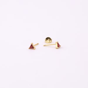 May include: Gold stud earrings featuring a triangular, red gemstone. The earrings are set against a white backdrop, with one earring post and a backing visible. The gemstones are a vivid red colour.