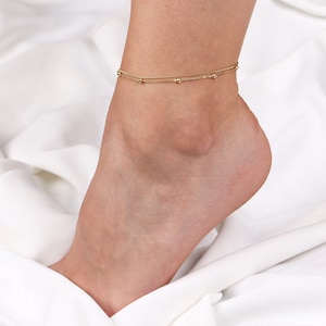 Peut inclure: Un bracelet de cheville en or avec deux chaînes et de petites perles. Le bracelet de cheville est sur une surface blanche.