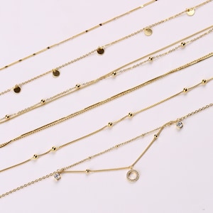 Peut inclure: Un ensemble de sept colliers en or avec différents styles de chaînes et des breloques. Certains colliers présentent de petits disques ronds, d'autres ont de petites perles et l'un a un petit pendentif rond avec un cristal transparent.