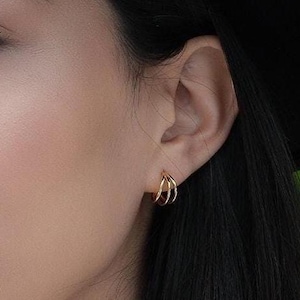 May include: A small gold hoop earring featuring a triple-band design. The earring consists of three thin, interconnected gold wires that form a single circular hoop shape, worn in the earlobe. The metal has a polished, reflective gold finish.