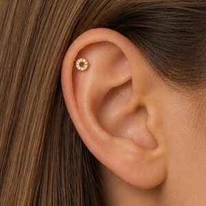 May include: A close-up of a person's ear with a small, gold-colored earring. The earring is circular, with a ring of small, clear stones around the edge. The earring is set in the upper cartilage of the ear.
