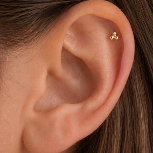 May include: A gold helix piercing featuring three small, round beads. The piercing is set on the upper cartilage of an ear, with brown hair in the background. The jewelry is a simple, elegant design.