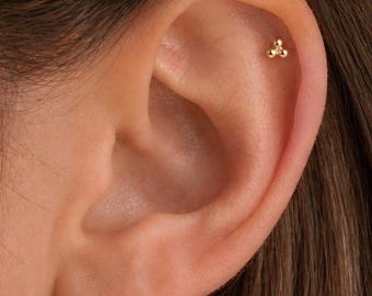 May include: A small gold stud earring featuring a cluster of three tiny spheres arranged in a triangular shape, positioned in the upper cartilage area of an ear. The earring has a polished metallic finish and a simple, compact design.