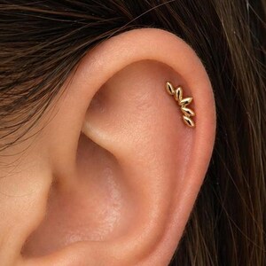 May include: A close-up of a person's ear adorned with gold jewelry. A small, delicate gold hoop earring is in the earlobe. Above, a gold helix piercing features a curved design of three connected leaf-like shapes.