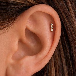 May include: A gold-toned cartilage earring featuring three small, clear, round-cut stones arranged in a vertical line. The earring is positioned in the upper ear, showcasing a minimalist design with a polished metal setting and secure backing.