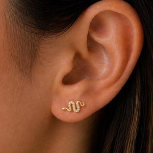 May include: A close-up of an ear with a gold snake stud earring. The earring is a small, delicate snake shape with a textured surface. The earring is set with small, sparkling stones.