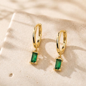 Peut inclure: Une paire de boucles d'oreilles créoles dorées, chacune dotée d'une petite breloque rectangulaire en pierre précieuse vert émeraude taille baguette suspendue. Les boucles d'oreilles ont une finition métallique polie et un fermoir à charnière.