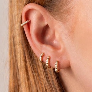 May include: A collection of gold-coloured earrings. The set features a cuff earring and three small hoop earrings, all embellished with tiny, sparkling stones. The earrings are shown on an ear, highlighting their design. The jewellery has a delicate and elegant appearance.