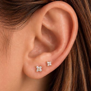 May include: Two gold stud earrings, each featuring a four-petaled flower design composed of small, clear, marquise-cut stones. The earrings are positioned on the earlobe, showcasing a delicate, symmetrical arrangement with a central gold setting.