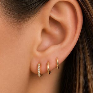 May include: Three gold hoop earrings, two small and one larger, are displayed on an ear. The largest hoop is encrusted with small, sparkling stones. The earrings are a classic jewelry item.
